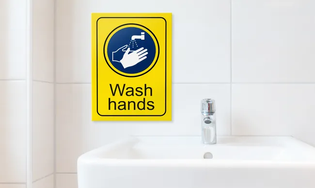 Trotec laser-engraved yellow Wash Hands safety sign above white sink in tiled bathroom