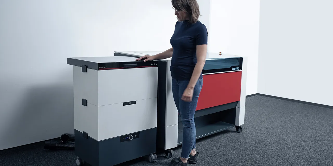 Woman operating Trotec Atmos Pure filtration system next to Speedy series CO₂ laser cutter
