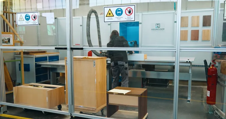 Operator using Trotec laser cutter in enclosed wood processing area with safety signs and furniture parts